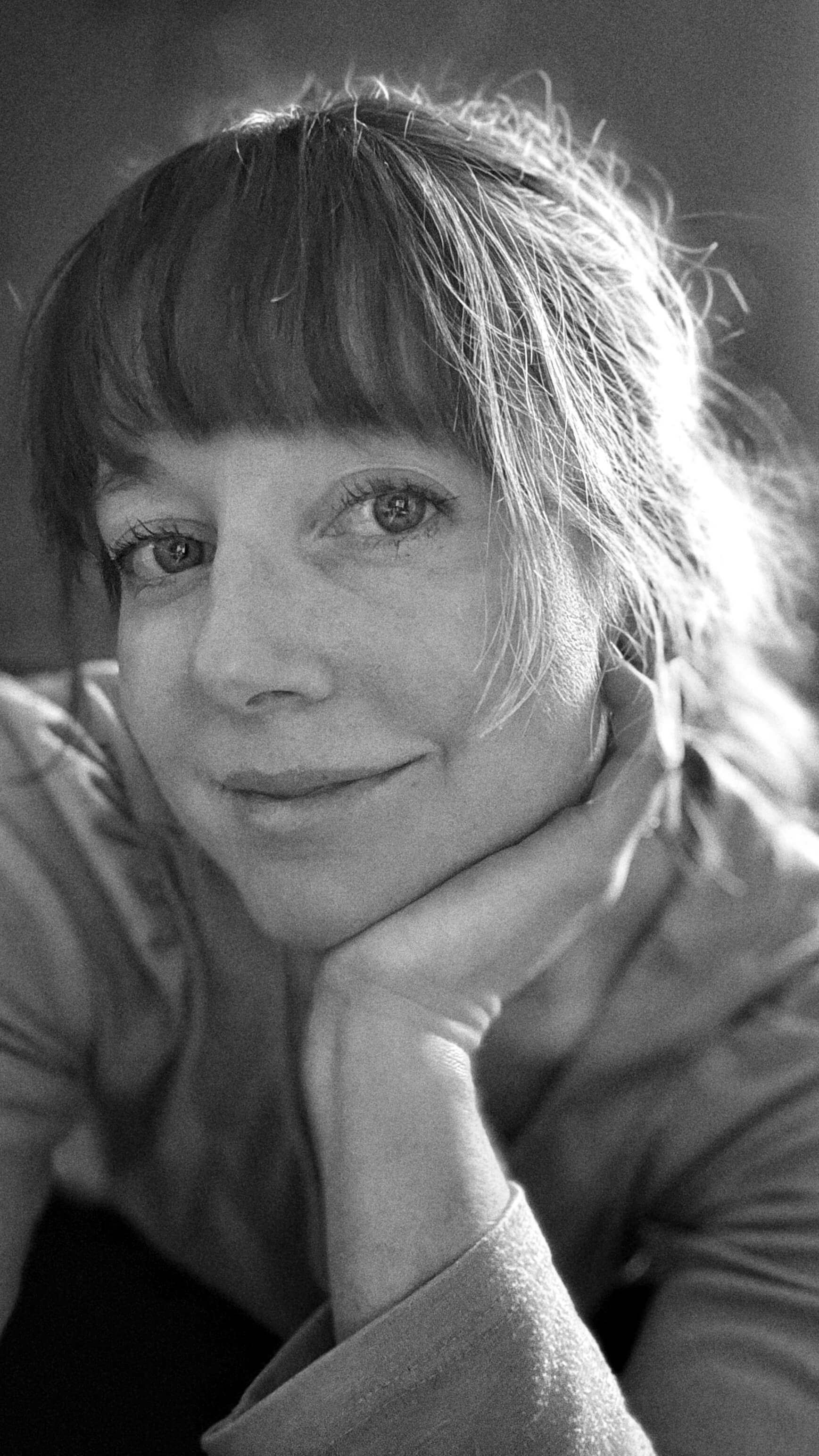 A black and white close-up of Clara Ritger looking directly at the camera with her head resting in her hand.