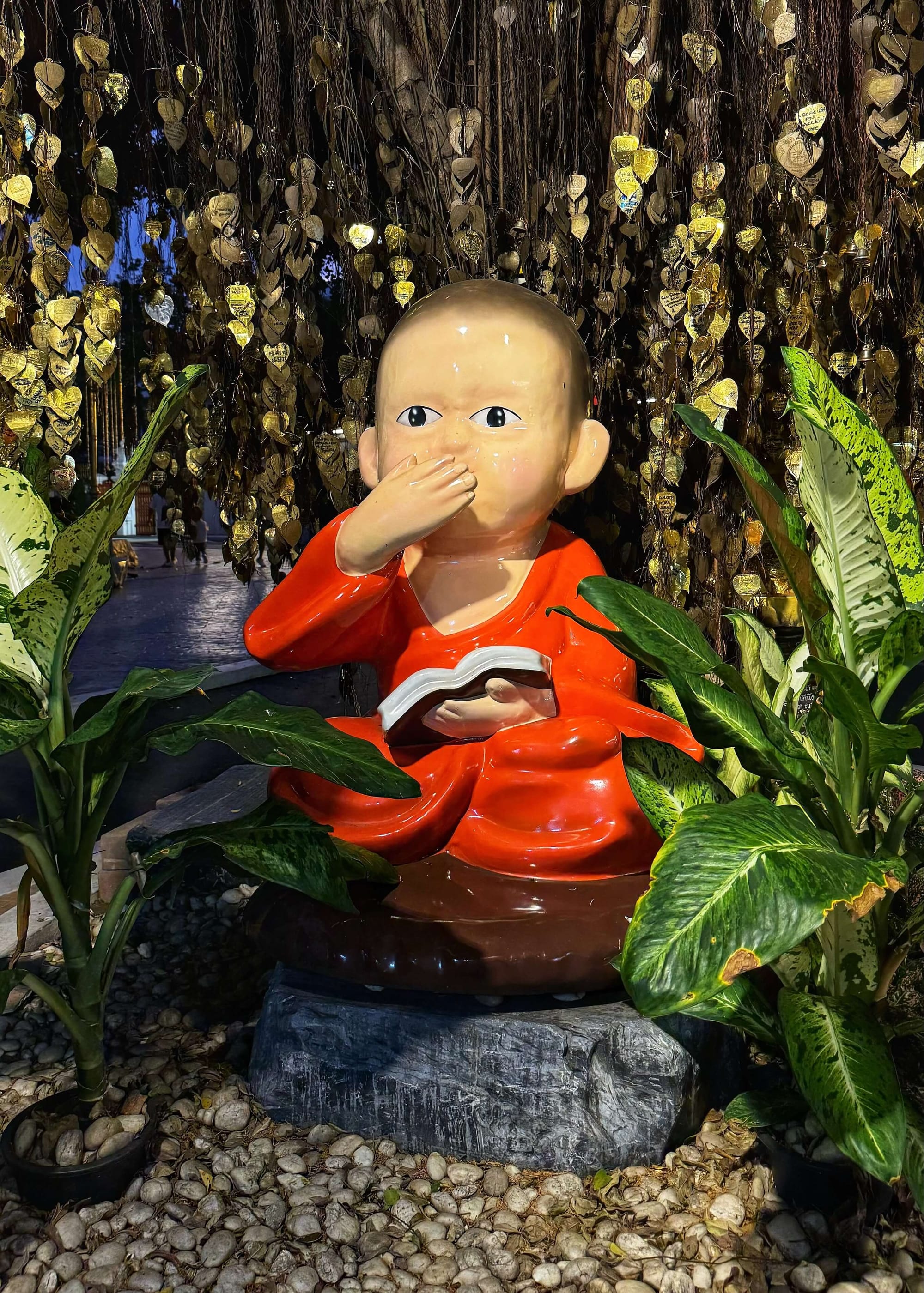 A bald, baby-like Buddha statue wearing an orange robe in front of a tree. 