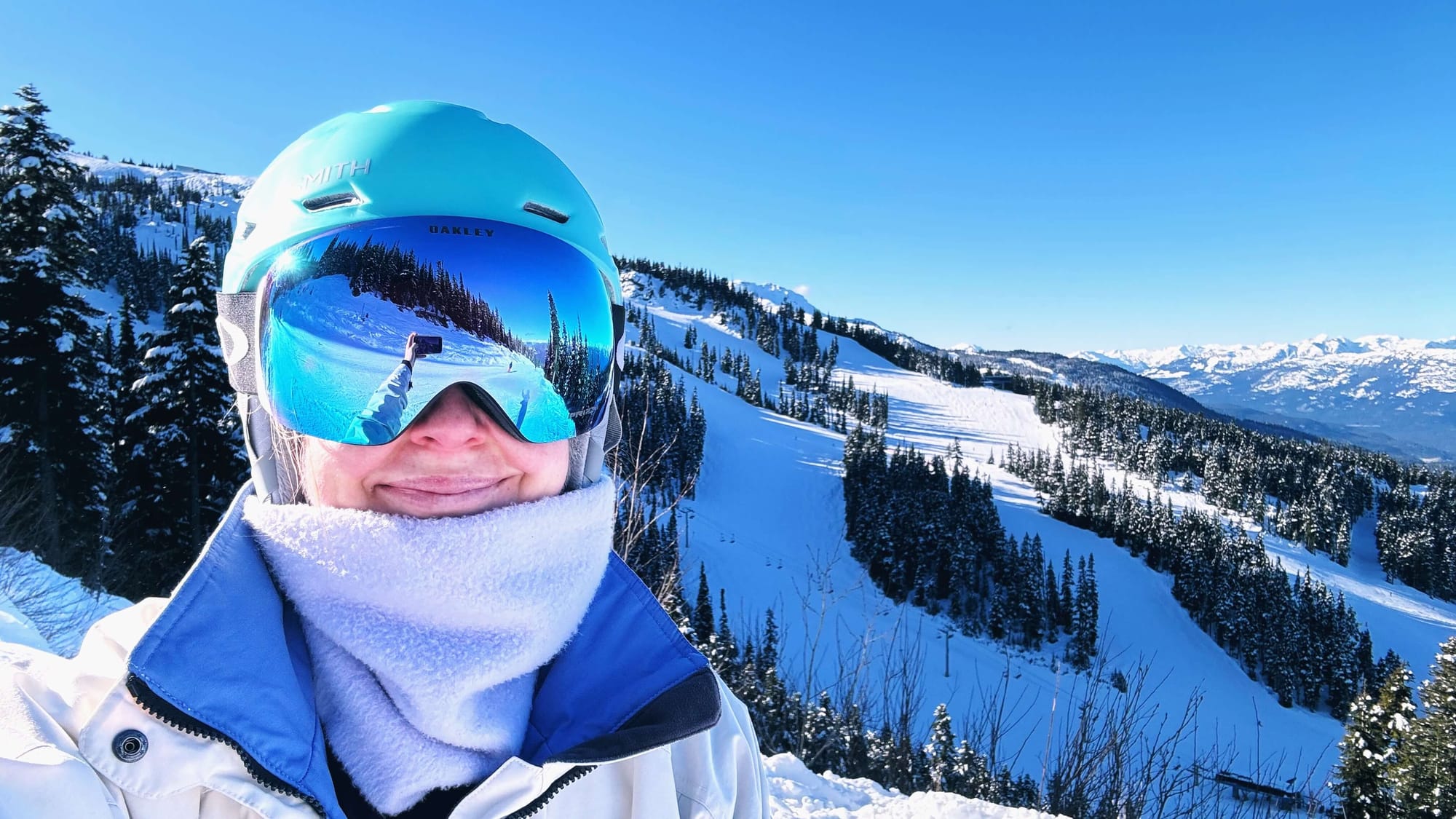 Clara Ritger poses for a selfie in a blue helmet and mirrored goggles, with three ski runs cutting through trees behind her.