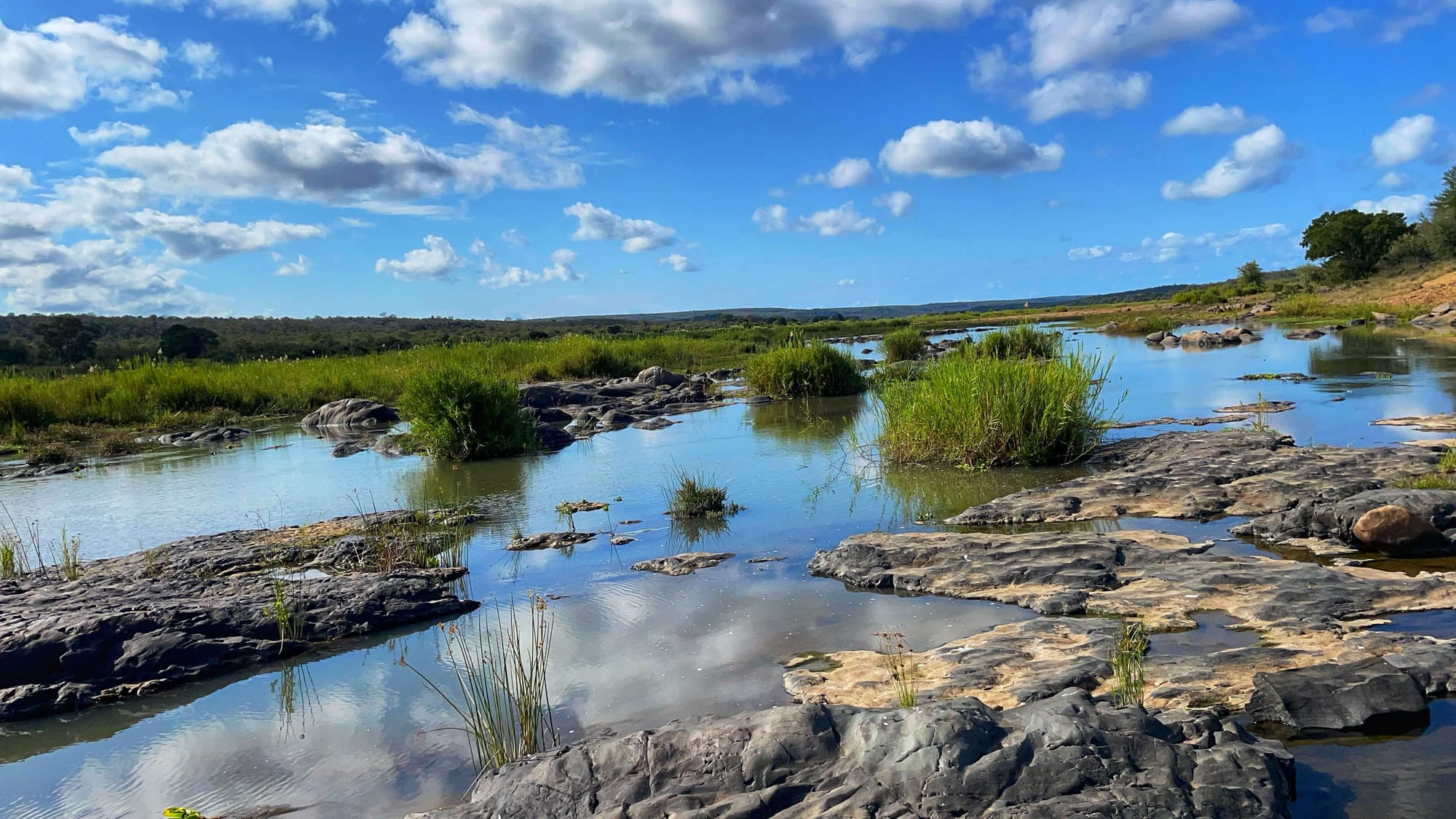 The blue sky and white clouds are reflected in the river. Rocky islands interrupt the flow, which is slow and smooth.