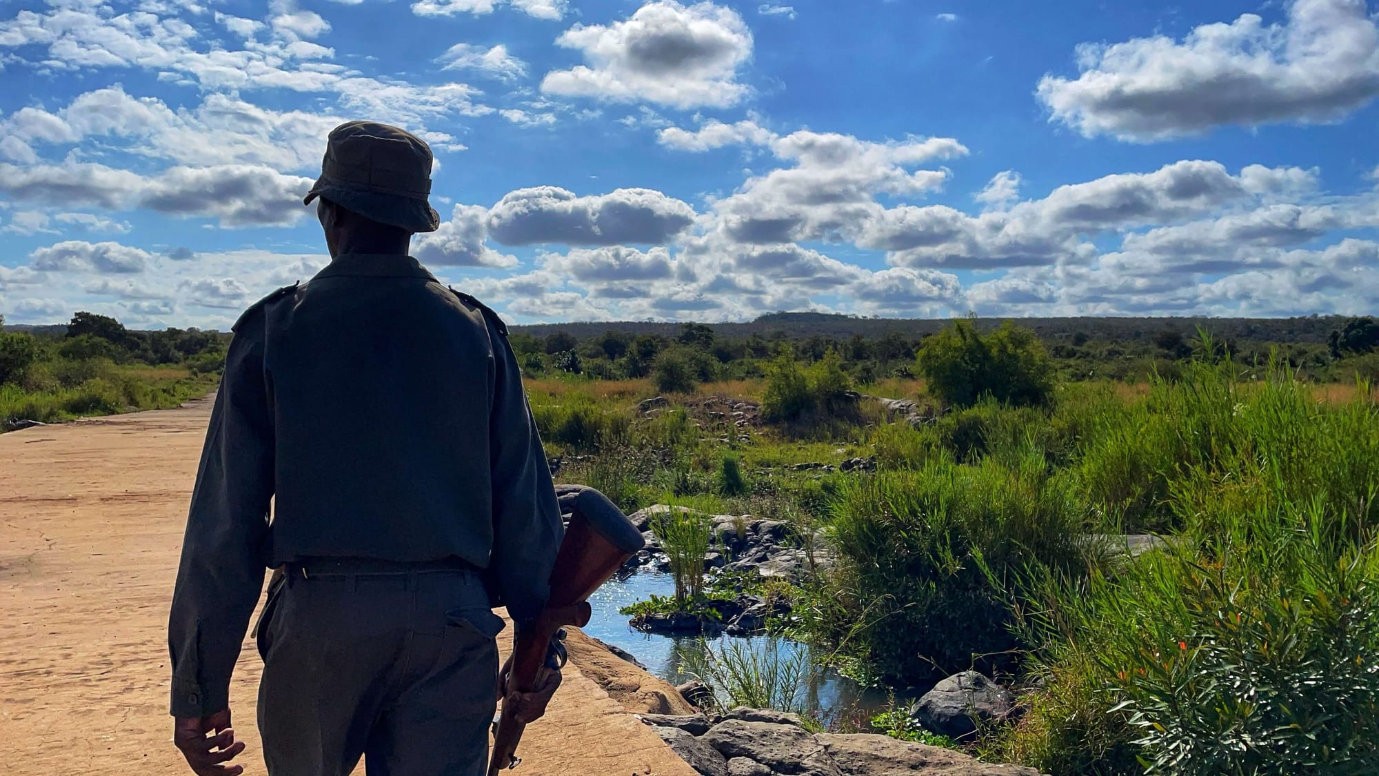 A man in uniform with a bucket hat and rifle walks along a yellow path. Around him is water and green bushes. The sky is blue and clouds are white.