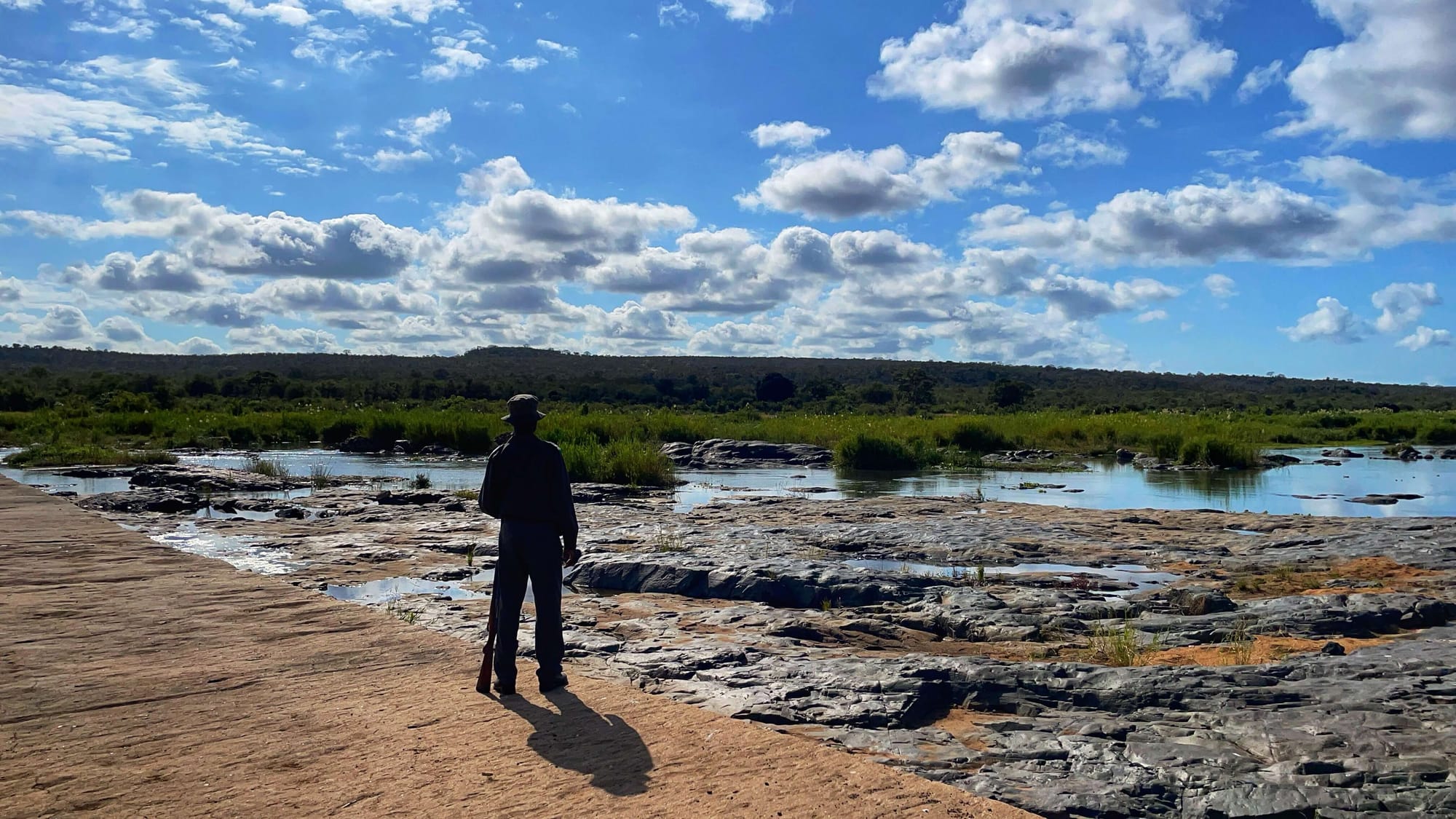 The silhouette of a ranger with a gun looking over a riverbed.