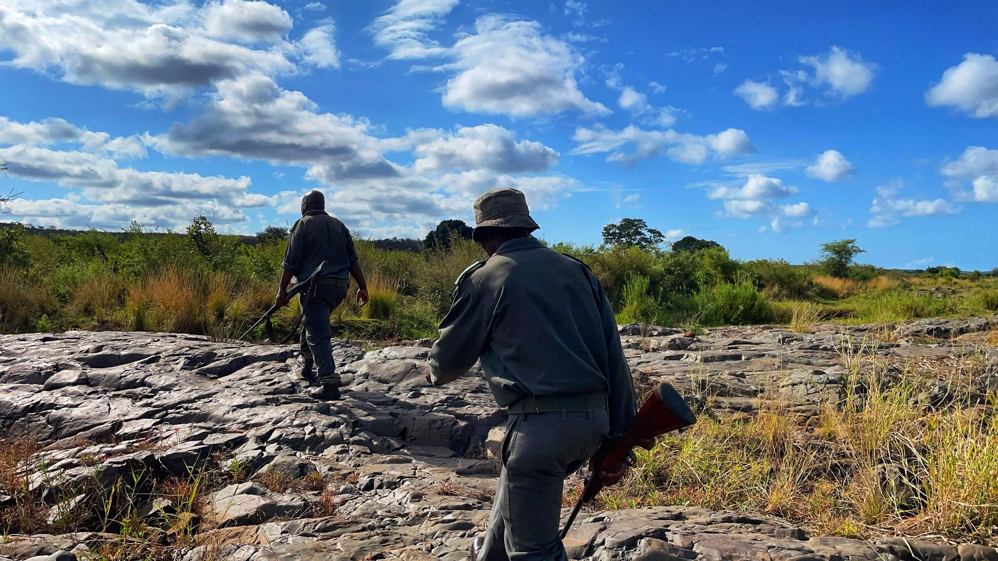 Two men in uniform with rifles walk up a rocky bank. The sky is blue and clouds are white.