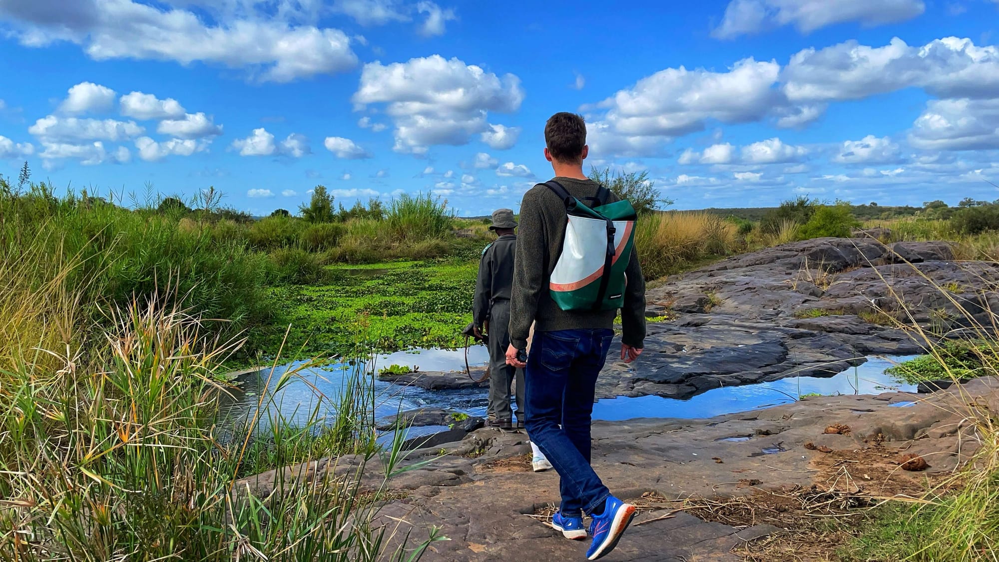 A young man with a backpack follows a man in uniform across the rocky waters. Green lily pads are visible in the distance.