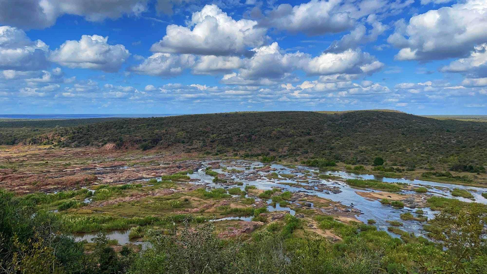 From a vantage point, pink and tan islands nearly cover the flow of the river. Another hill can be seen across the way.