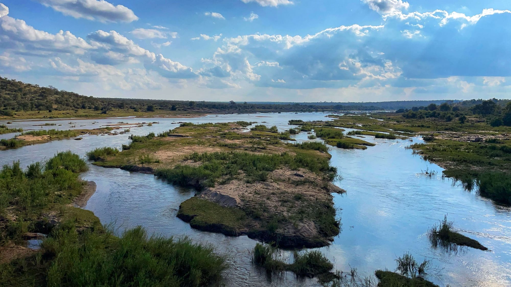 Patches of land interrupt a blue river, like islands. The river reflects the blue sky and white clouds.