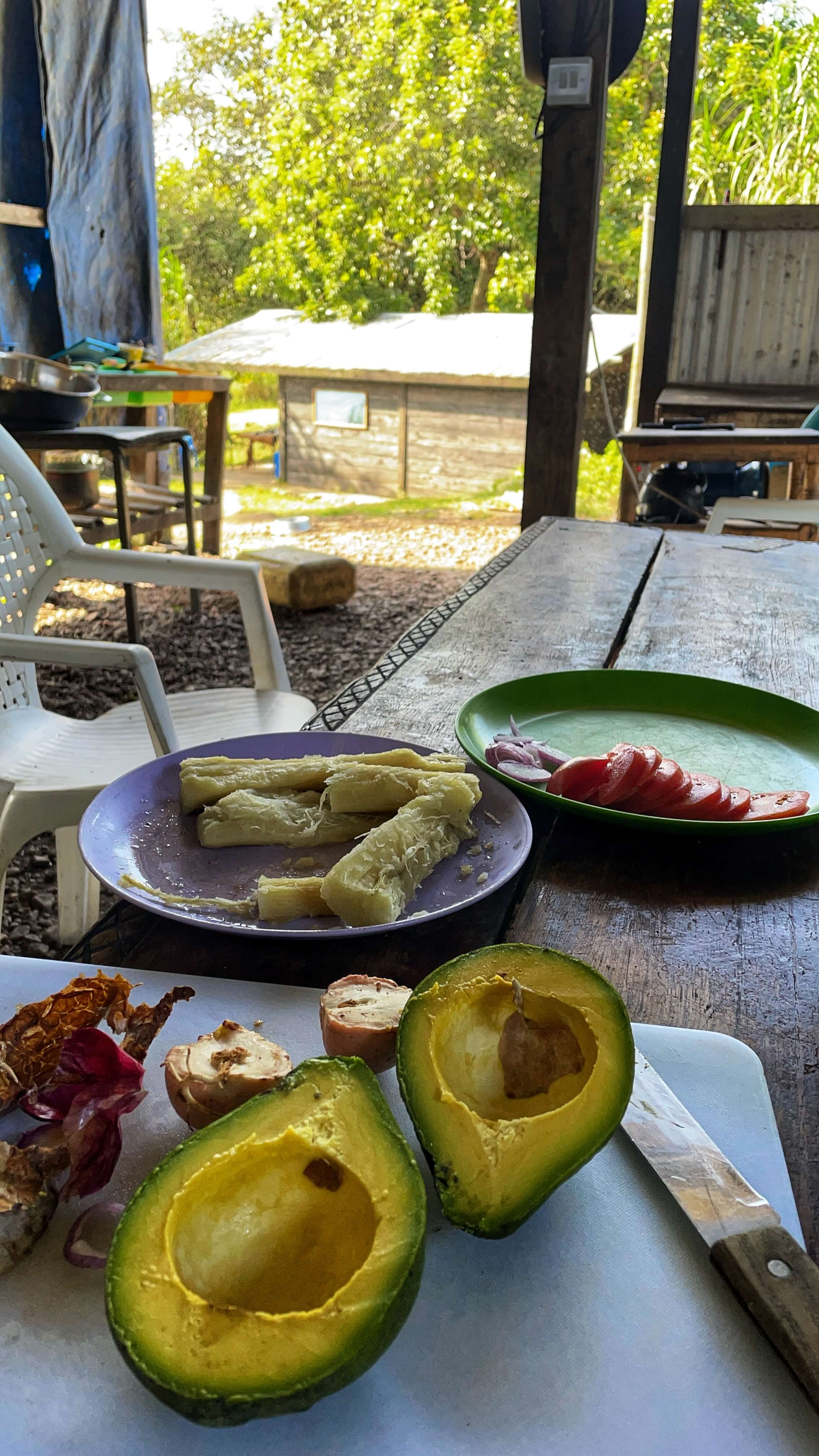 An avocado split in half, a plate of yellow cassava, and another plate of sliced tomatoes and onions.