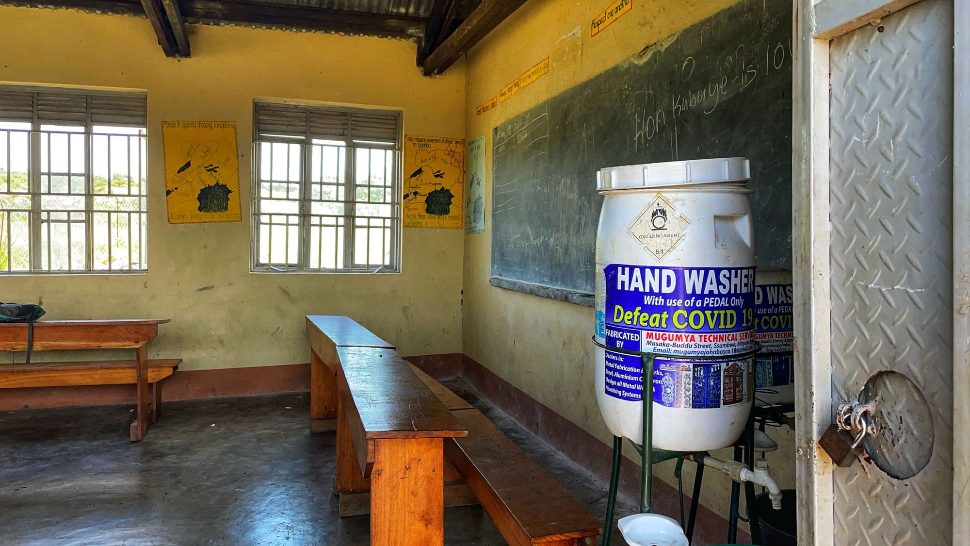 A big white tub that reads "HAND WASHER DEFEAT COVID" sits next to a blackboard in a yellow walled school room.