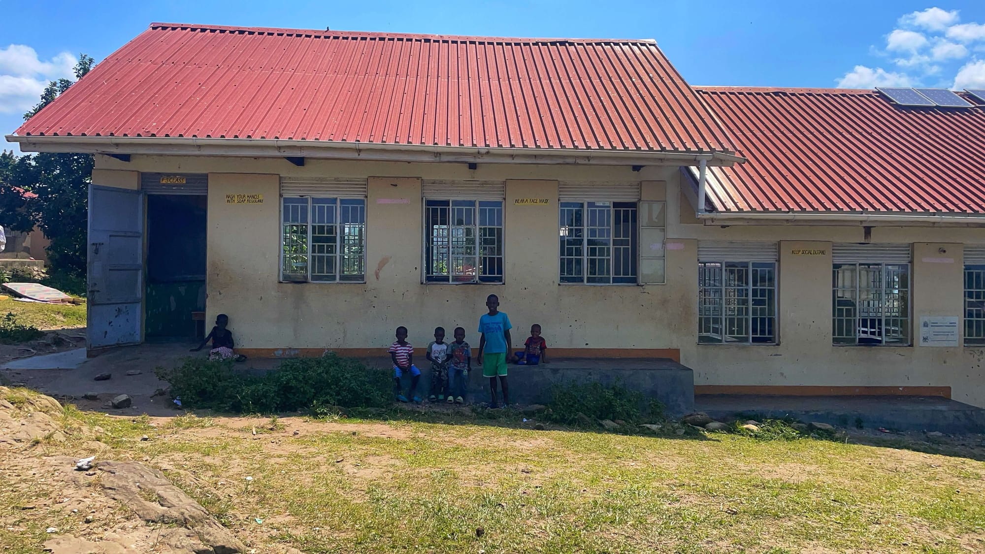 Six children wait outside of a yellow, one-story building with a red roof.