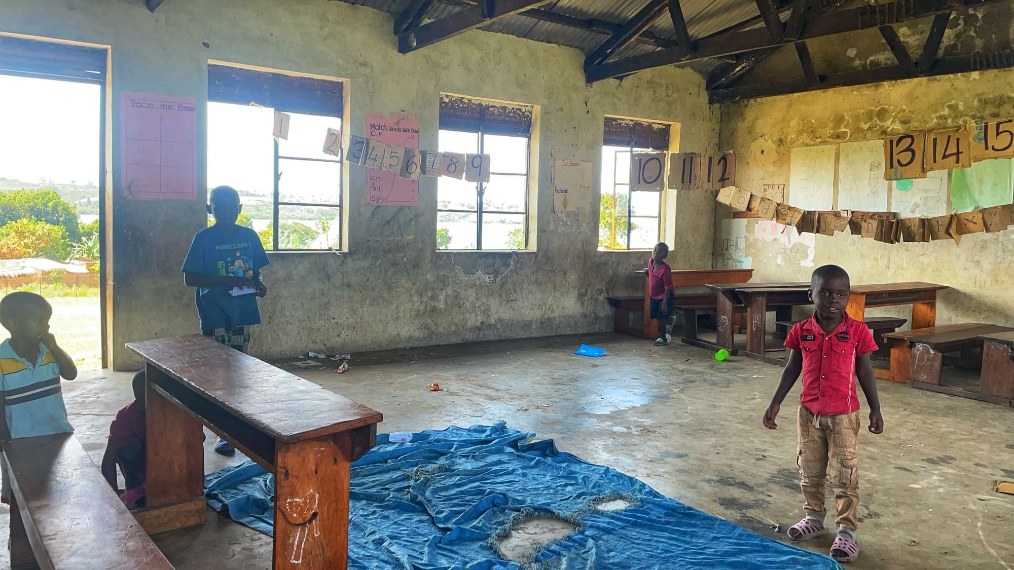 Three children stand in a simple classroom with concrete walls and numbers on brown paper hanging across the room.