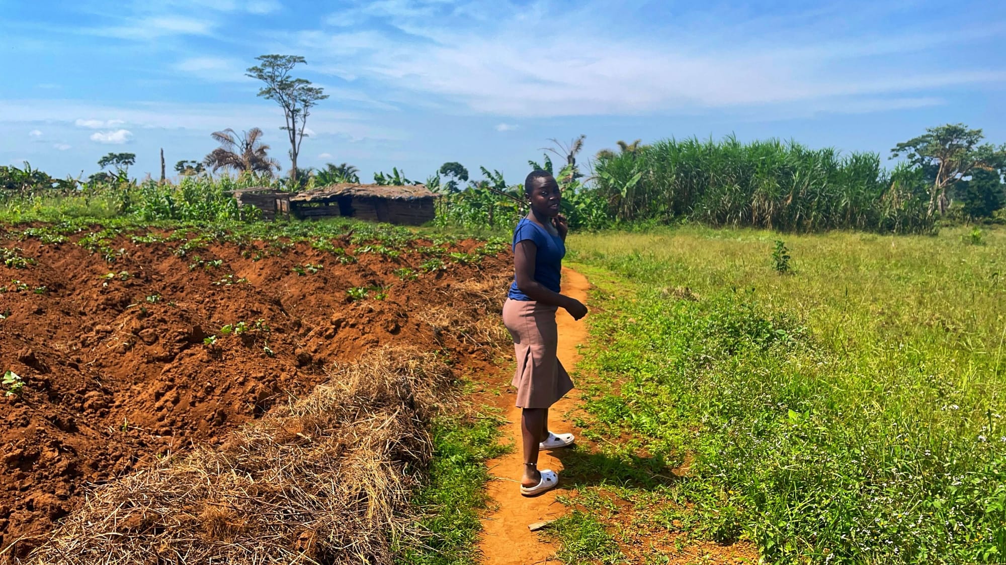 Sumi, a teenage Ugandan girl, looks at the camera. On her left are dirt mounds with freshly planted crops. On her right is a green grassy meadow.