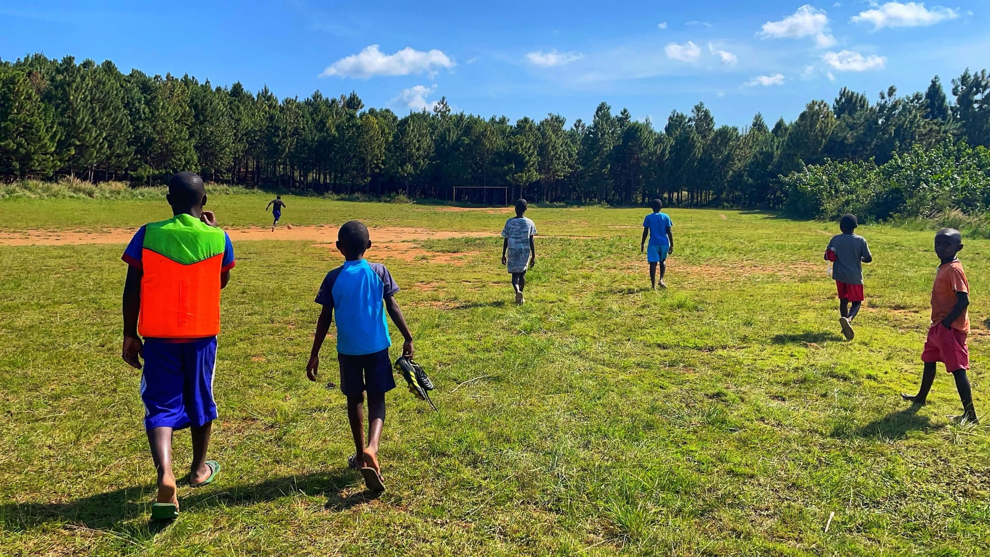 Boys in colorful clothes gather on a grassy field. In the distance, one runs with a soccer ball.