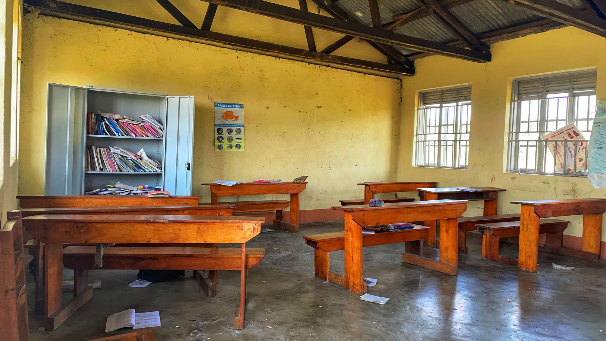 A yellow walled room with brown wooden benches as desks. A metal storage cabinet with open doors reveals messy books inside.